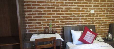 Una habitación con pared de ladrillo, un escritorio de madera, una silla, una cama con ropa blanca y una almohada roja, y un pequeño estante con una botella y vasos.
