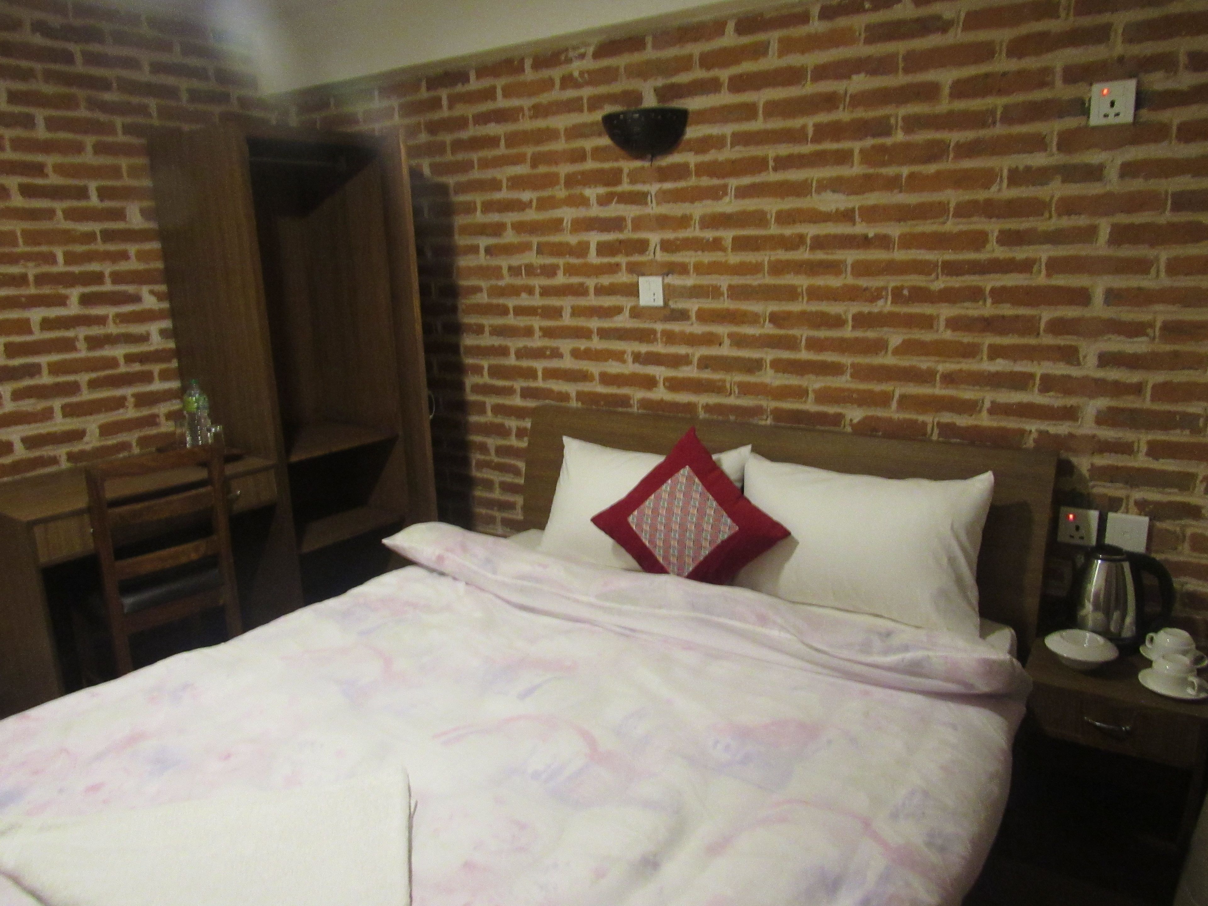 A hotel room with a brick wall, a wooden wardrobe, a bed with a floral bedspread, a red pillow, and a bedside table with a kettle and cups.