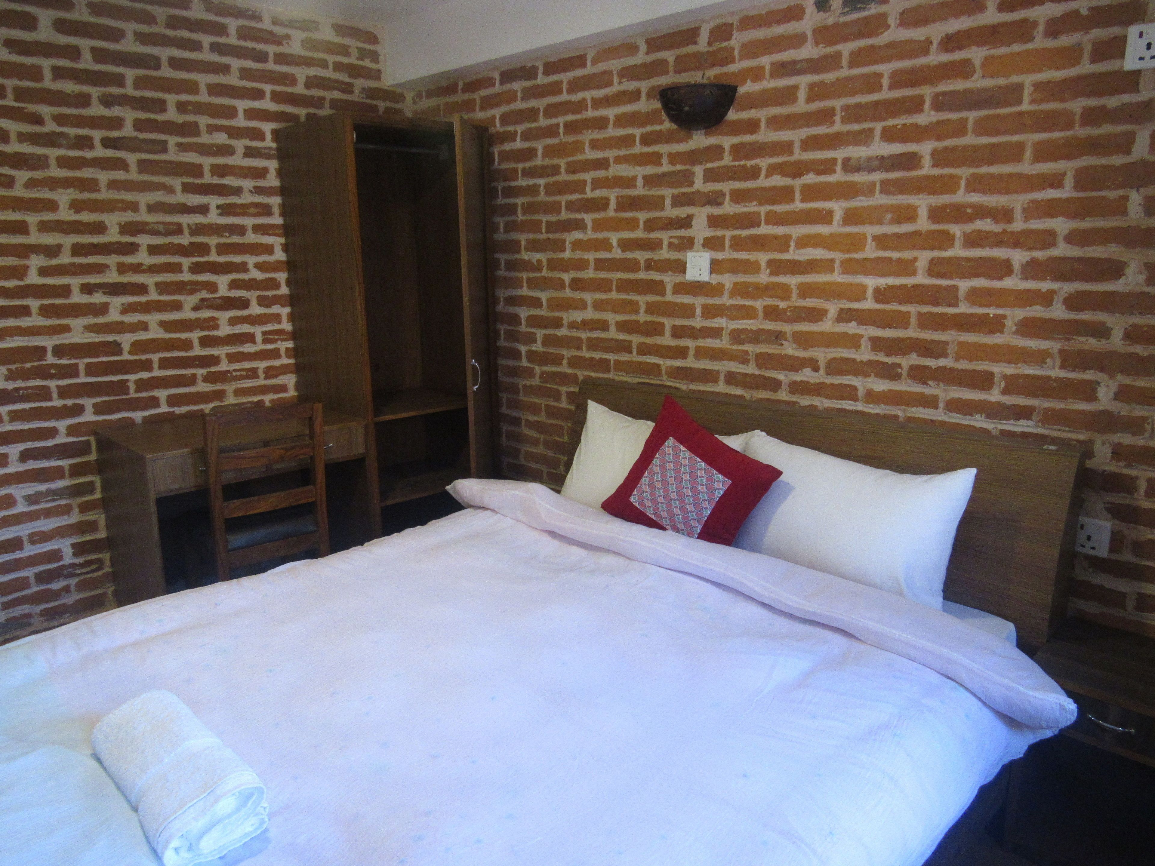 Una habitación con pared de ladrillo, una cama de madera con almohadas blancas y rojas, un armario de madera y una pequeña silla de madera.