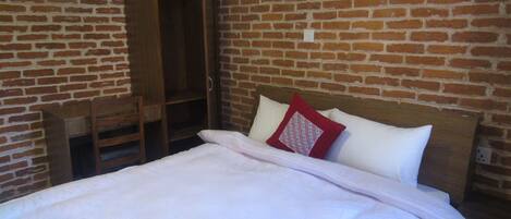 Una habitación con pared de ladrillo, una cama de madera con almohadas blancas y rojas, un armario de madera y una pequeña silla de madera.