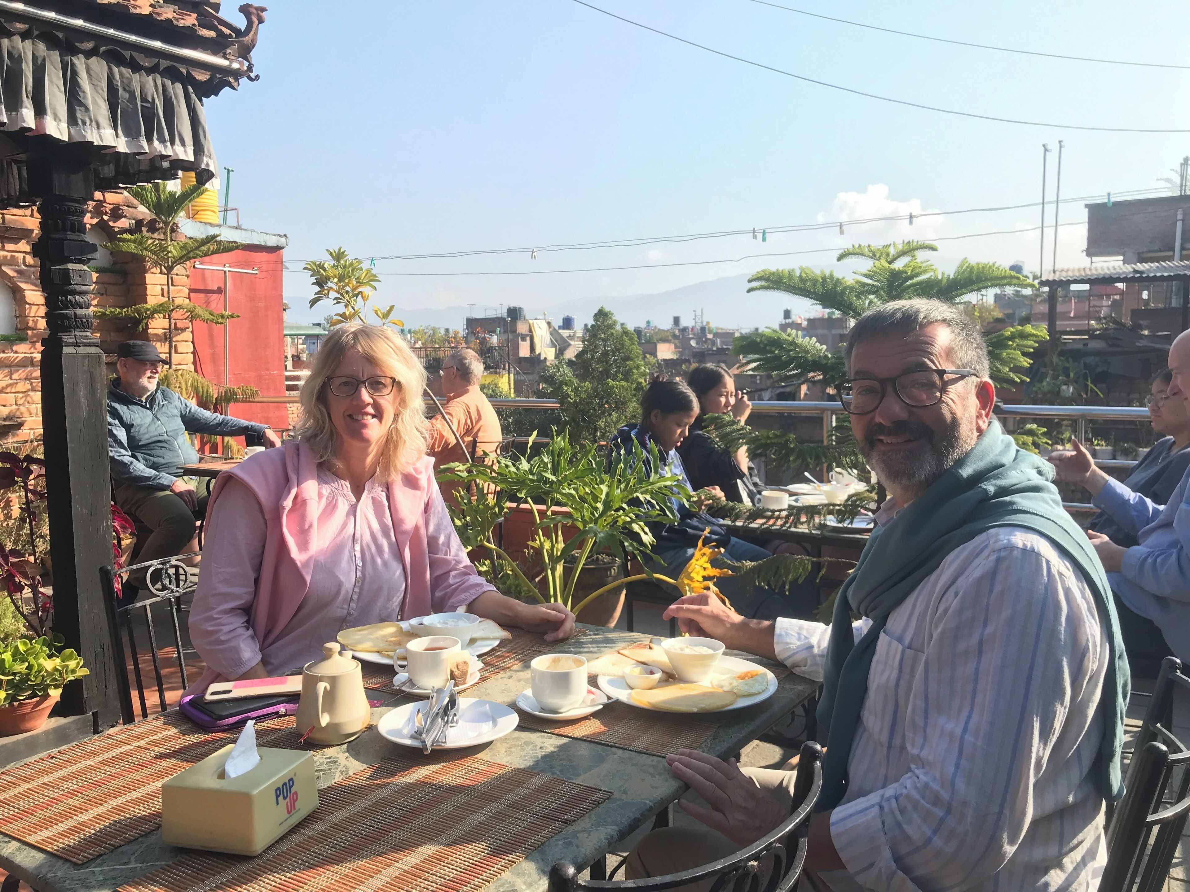 A group of people dining outdoors with a table set for tea and light snacks, surrounded by greenery and buildings in the background.