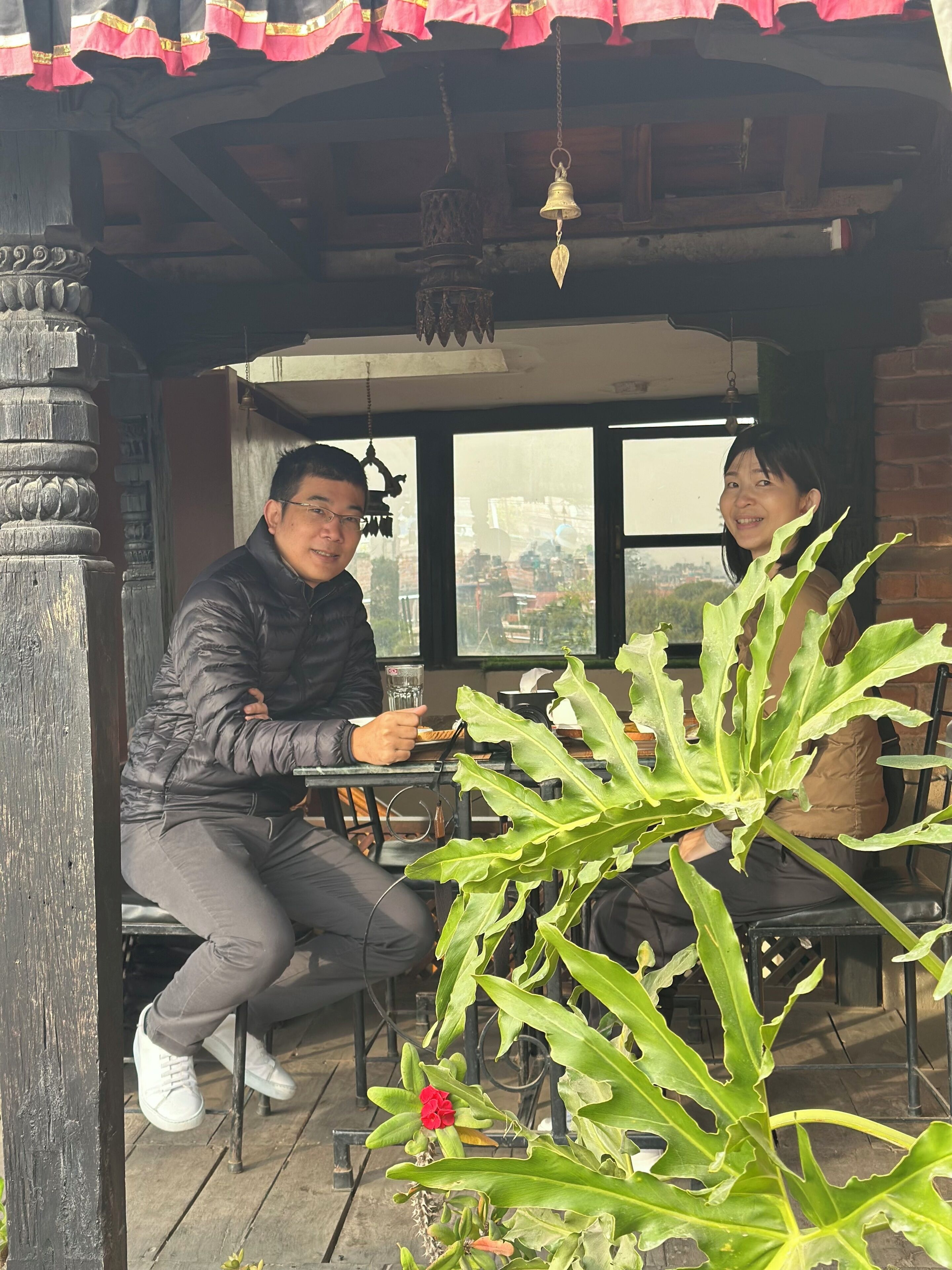 Un hombre y una mujer sentados a una mesa en un espacio al aire libre techado, con vistas a la ciudad a través de amplios ventanales.