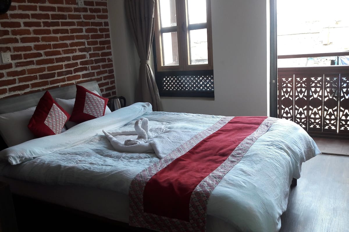 A hotel room with a brick wall, a bed with white bedding and red pillows, a window with a view, and a balcony with a lattice design.