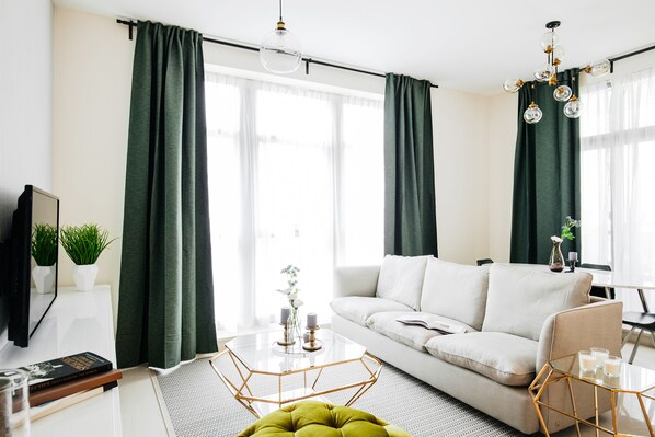 A modern living room with a white sofa, green curtains, a glass coffee table, and a green ottoman.
