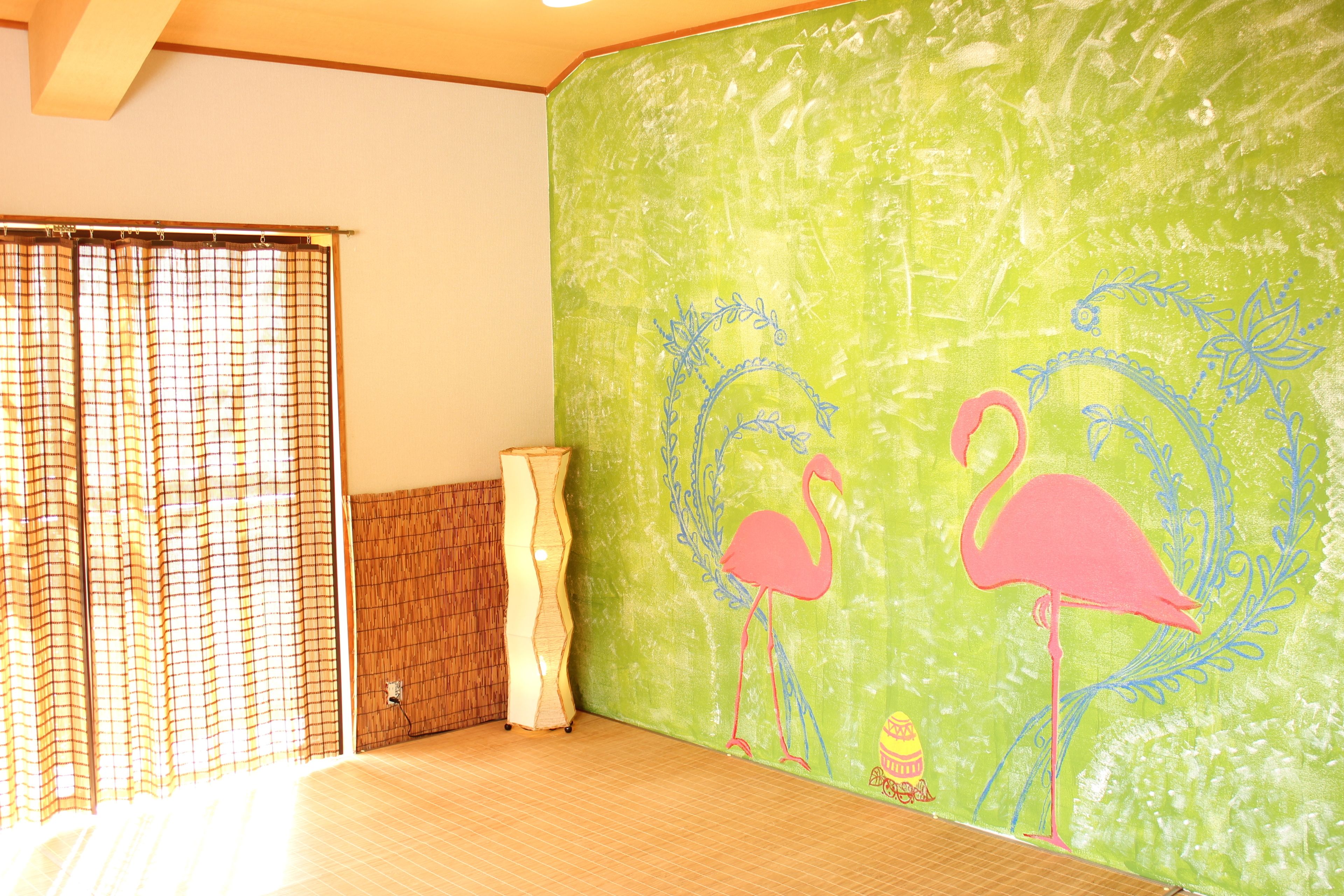 A room with a green wall, a bamboo mat, a white vase, and two pink flamingo drawings.