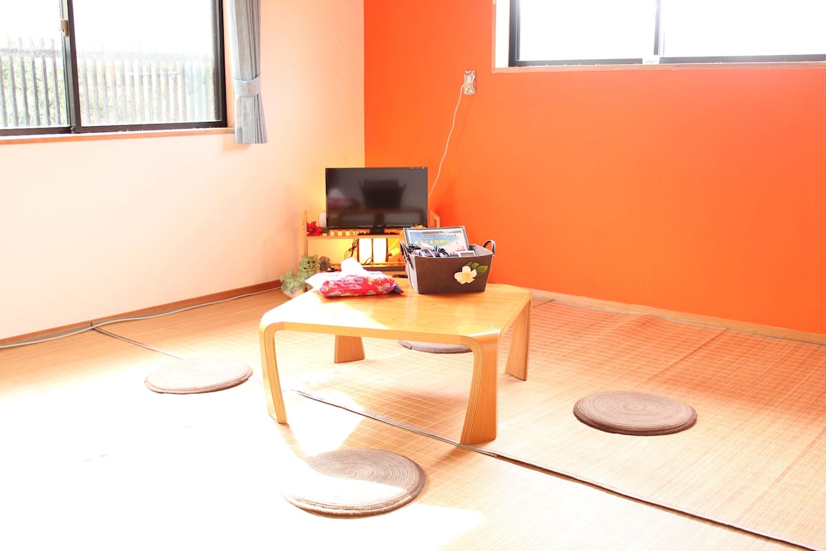 Une pièce avec un sol recouvert de tatamis, une télévision, une petite table avec un panier, et une fenêtre avec des rideaux.