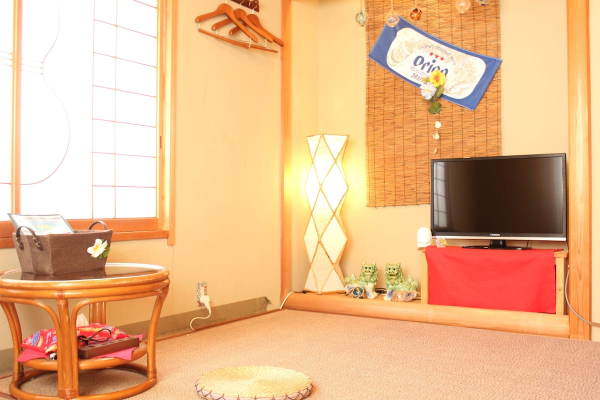 Une pièce comprenant une télévision, un fauteuil en osier, une petite table avec un sac, un tissu rouge et un objet décoratif accroché au mur.