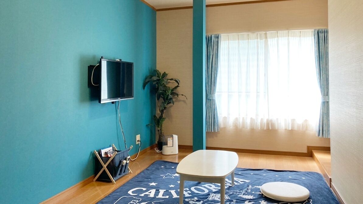 Una habitación con paredes verde azulado, un televisor montado, una planta, una ventana con cortinas, una alfombra azul con el texto 'CALIFORNIA', una silla blanca y una pequeña mesa redonda.