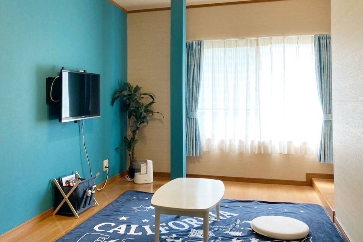 A room with teal walls, a mounted TV, a plant, a window with curtains, a blue rug with 'CALIFORNIA' text, a white chair, and a small round table.