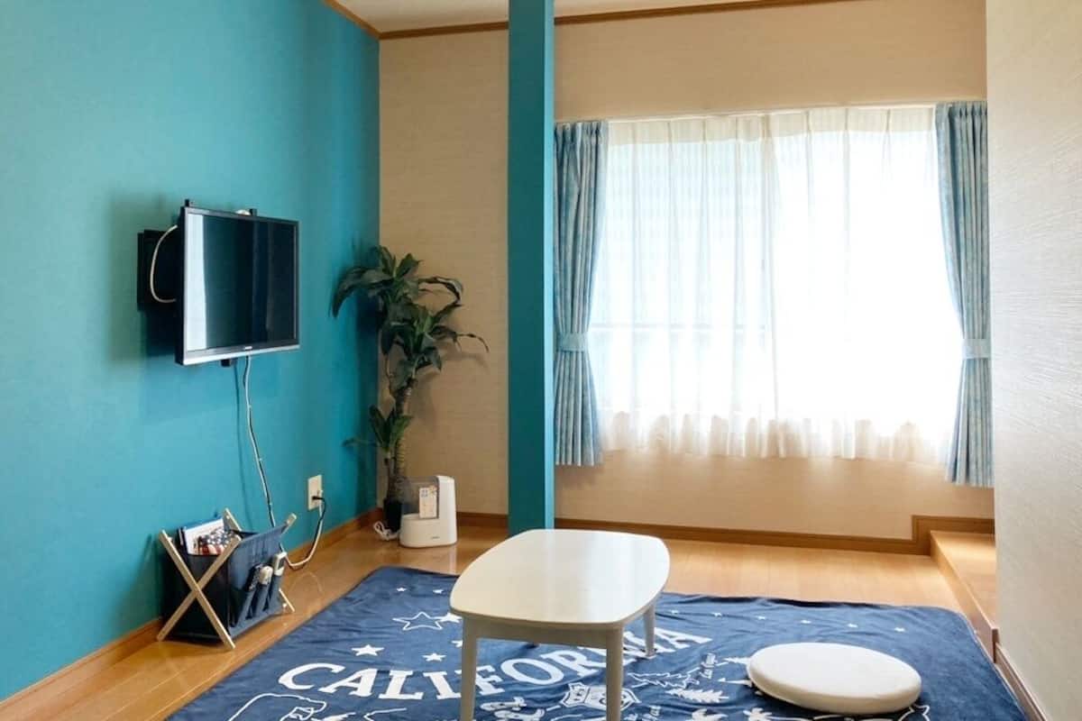 Una habitación con paredes verde azulado, un televisor montado, una planta, una ventana con cortinas, una alfombra azul con el texto 'CALIFORNIA', una silla blanca y una pequeña mesa redonda.