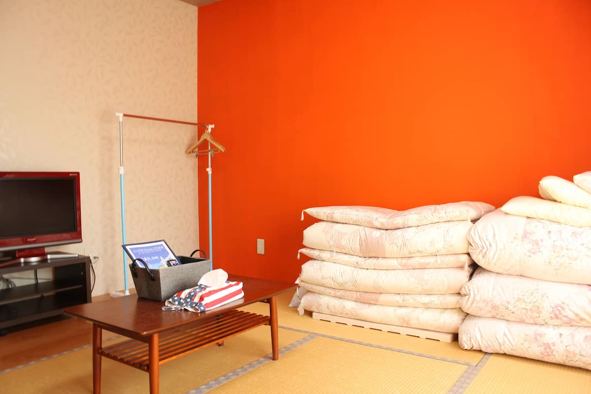 A room with a television, a table with a laptop and a bag, and a stack of pillows against an orange wall.
