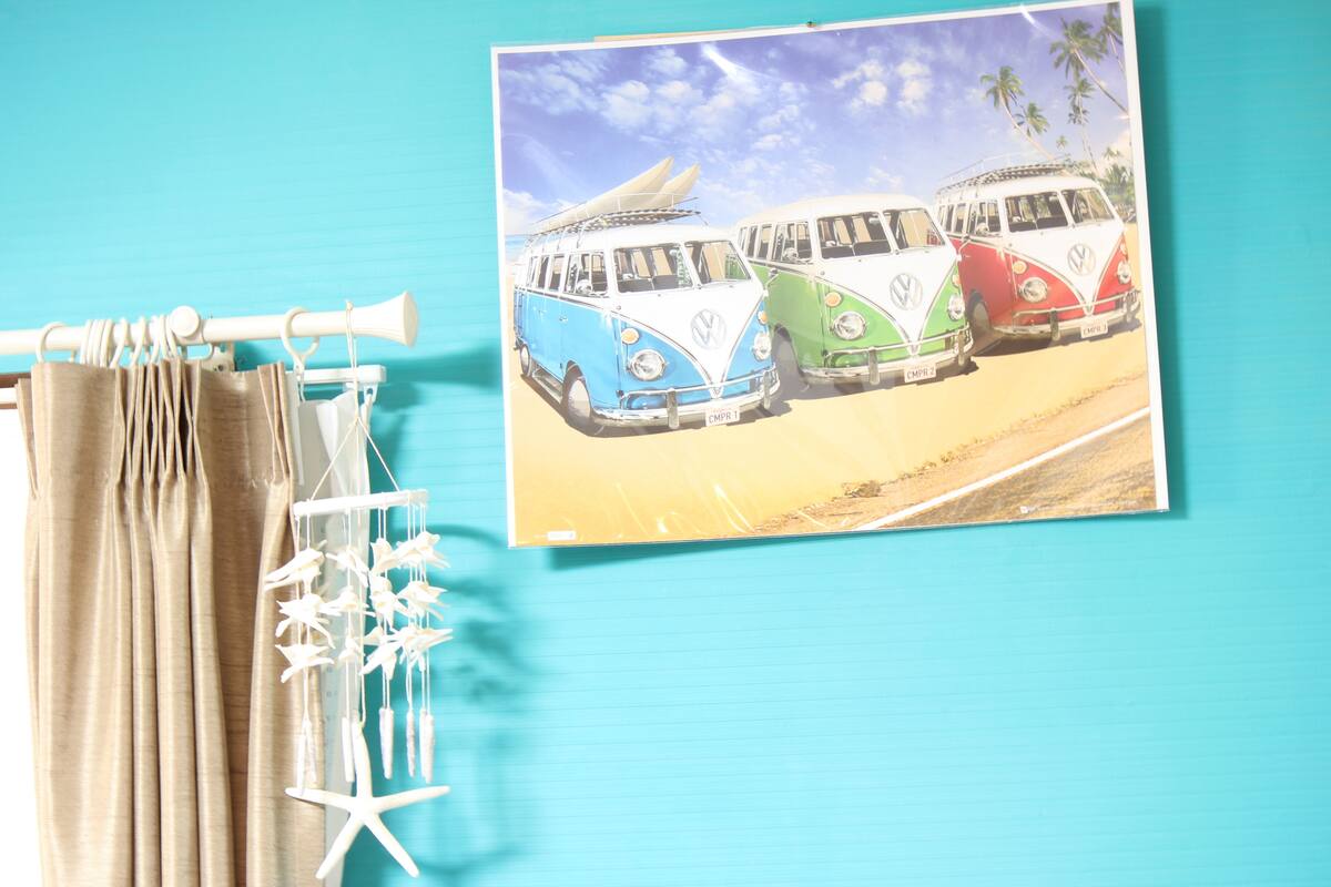 A room with a teal wall, a framed picture of vintage VW vans, a curtain rod with beige curtains, and a white starfish decoration.