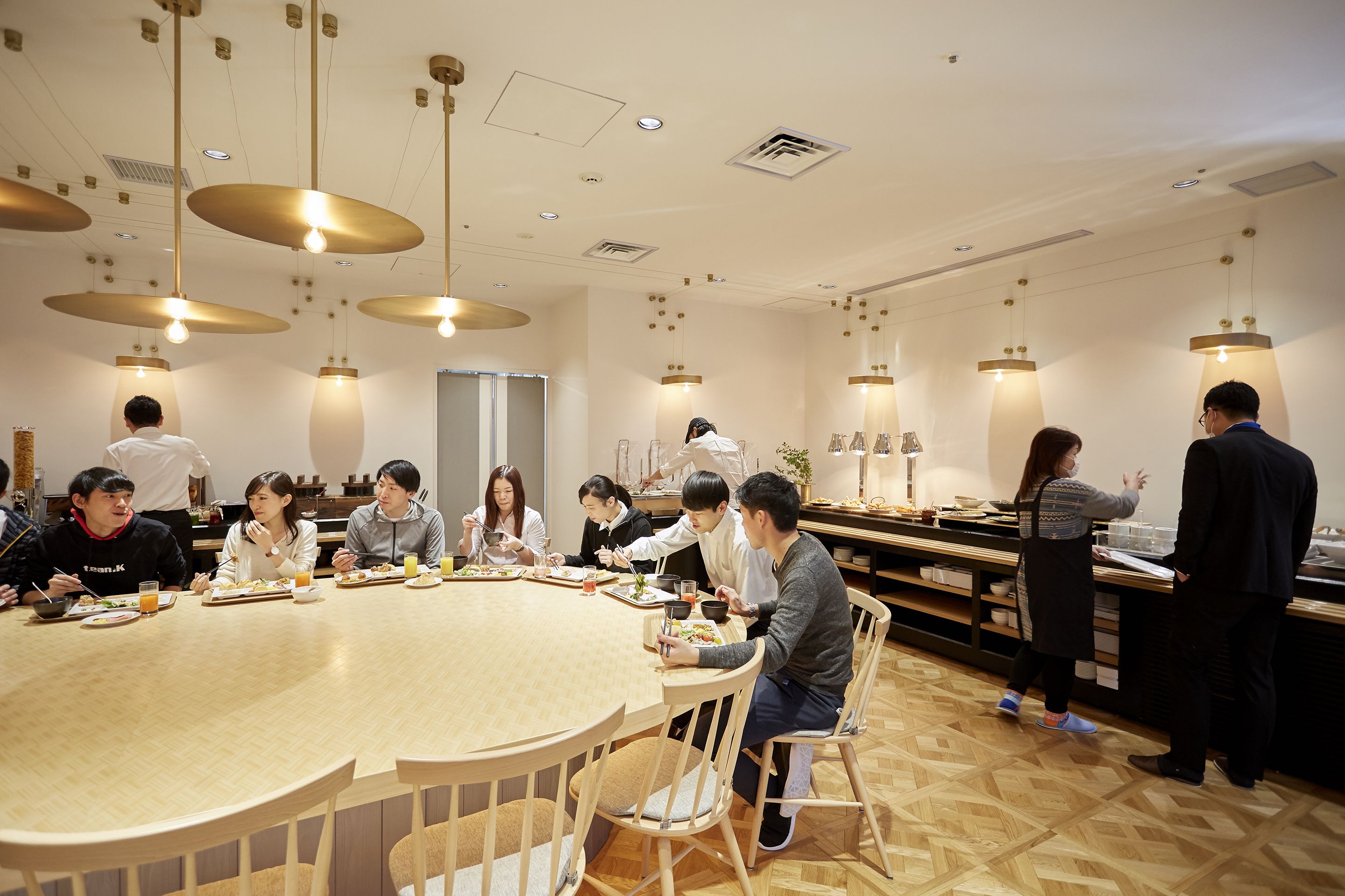 Un espace repas avec une table ronde en bois entourée de chaises, un comptoir de cuisine où un chef est en train de travailler, et des luminaires suspendus.