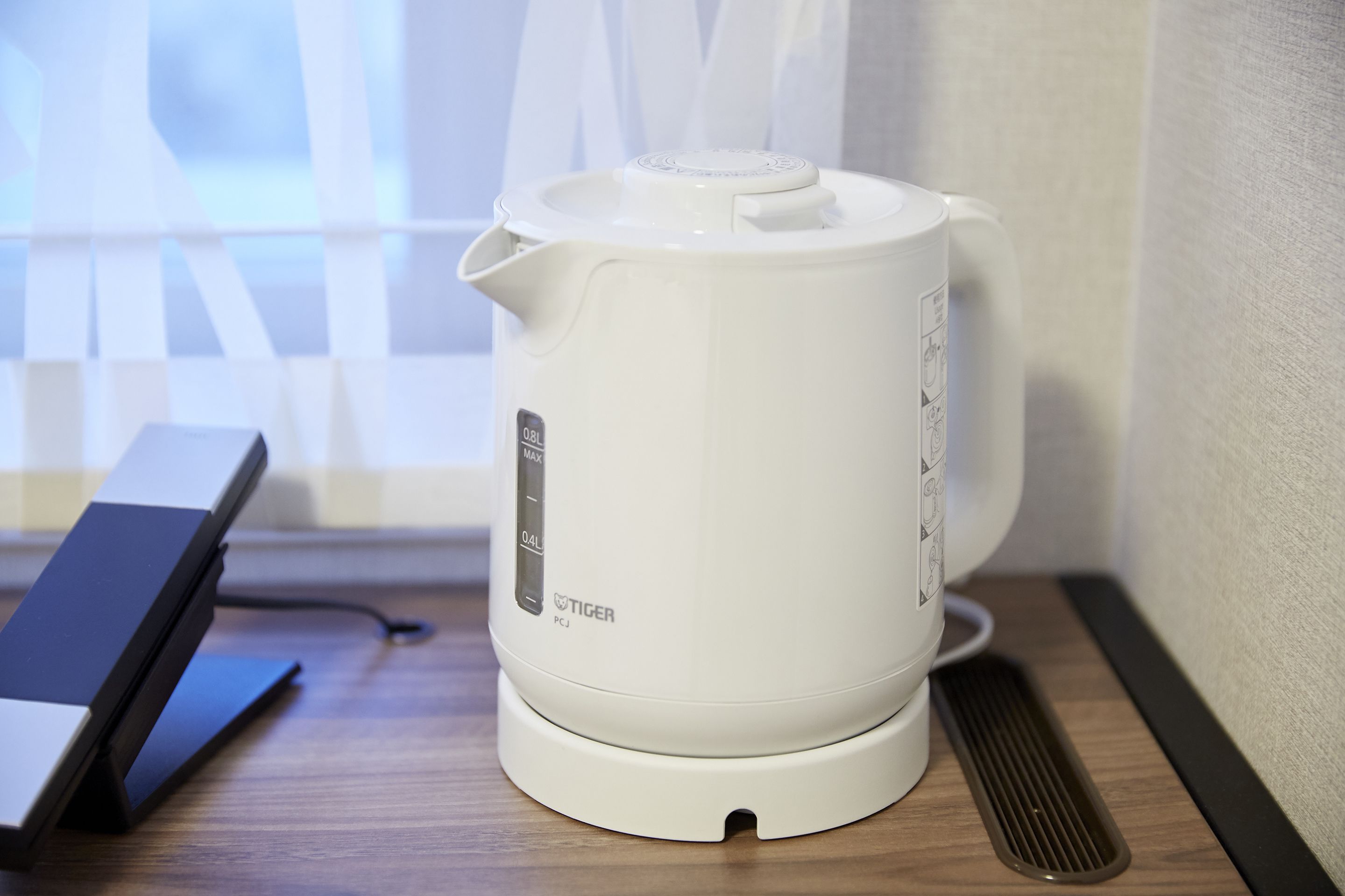 A white electric kettle on a wooden table next to a black device, possibly a charger, with a window in the background.