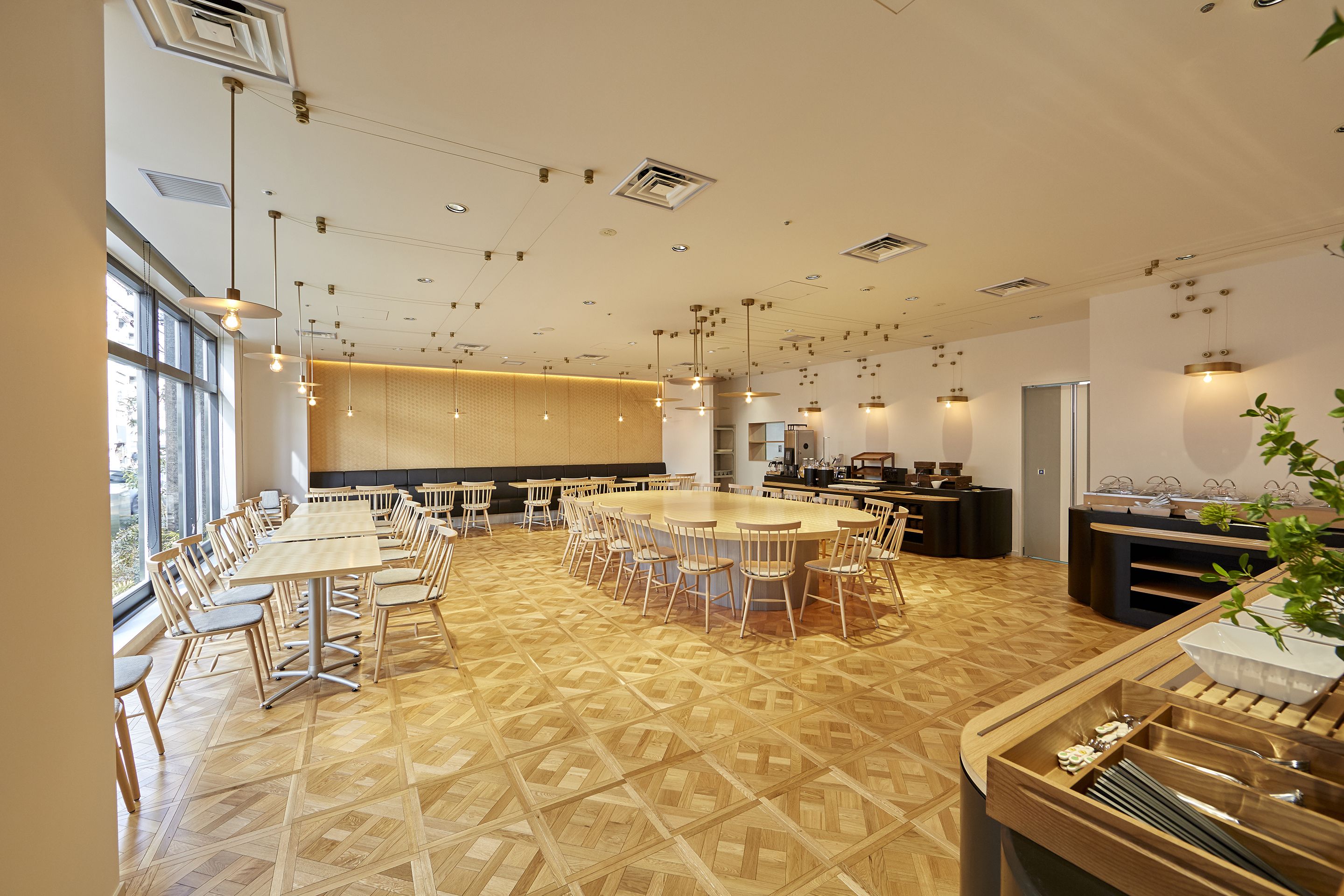 A modern dining area with a large central table surrounded by chairs, a kitchen area with a buffet, and large windows providing natural light.