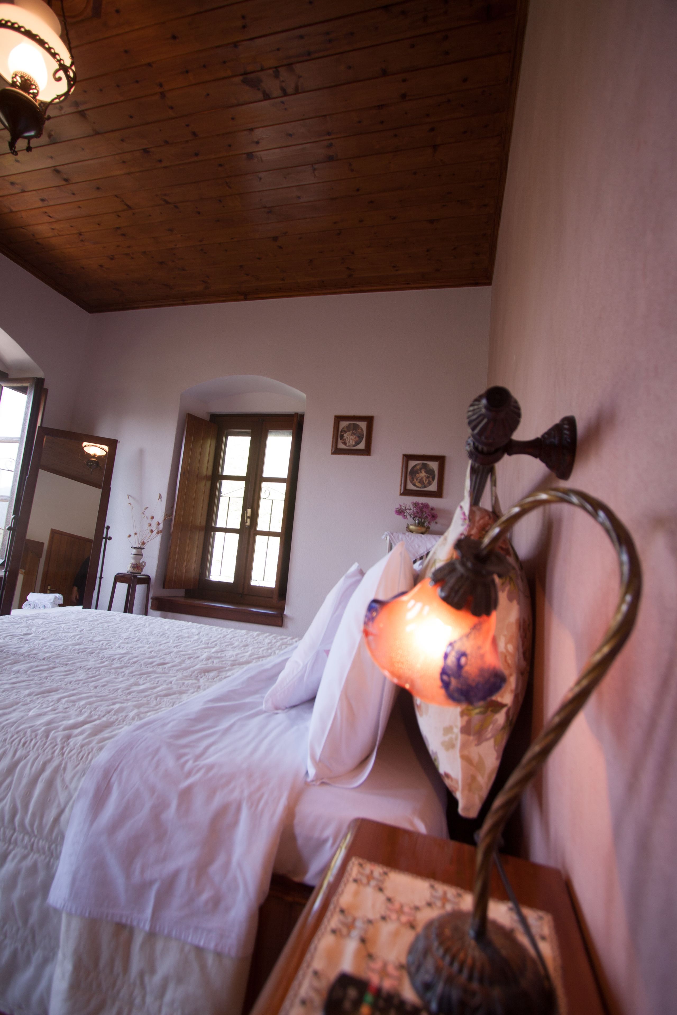 A bedroom with a wooden ceiling, a bed with white bedding, a wall-mounted lamp, and a window with curtains.