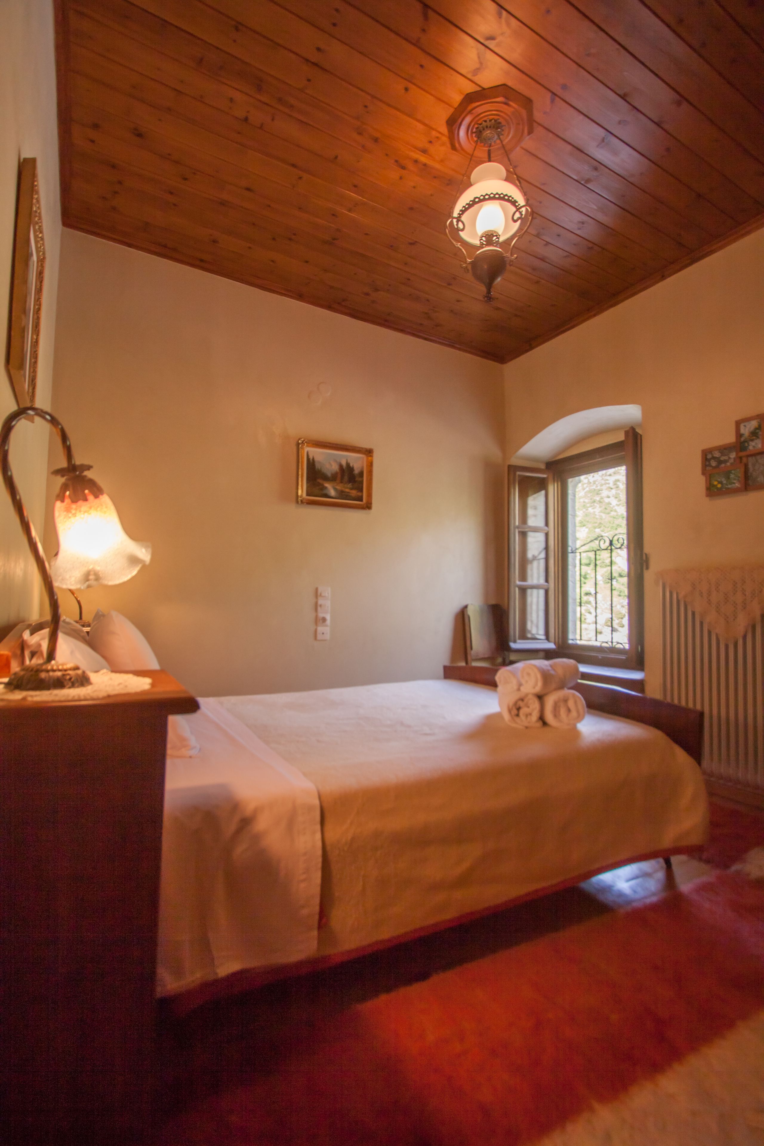 Un dormitorio con techo de madera, una cama con colcha blanca, una lámpara de noche, una ventana con vistas a la vegetación y un radiador.