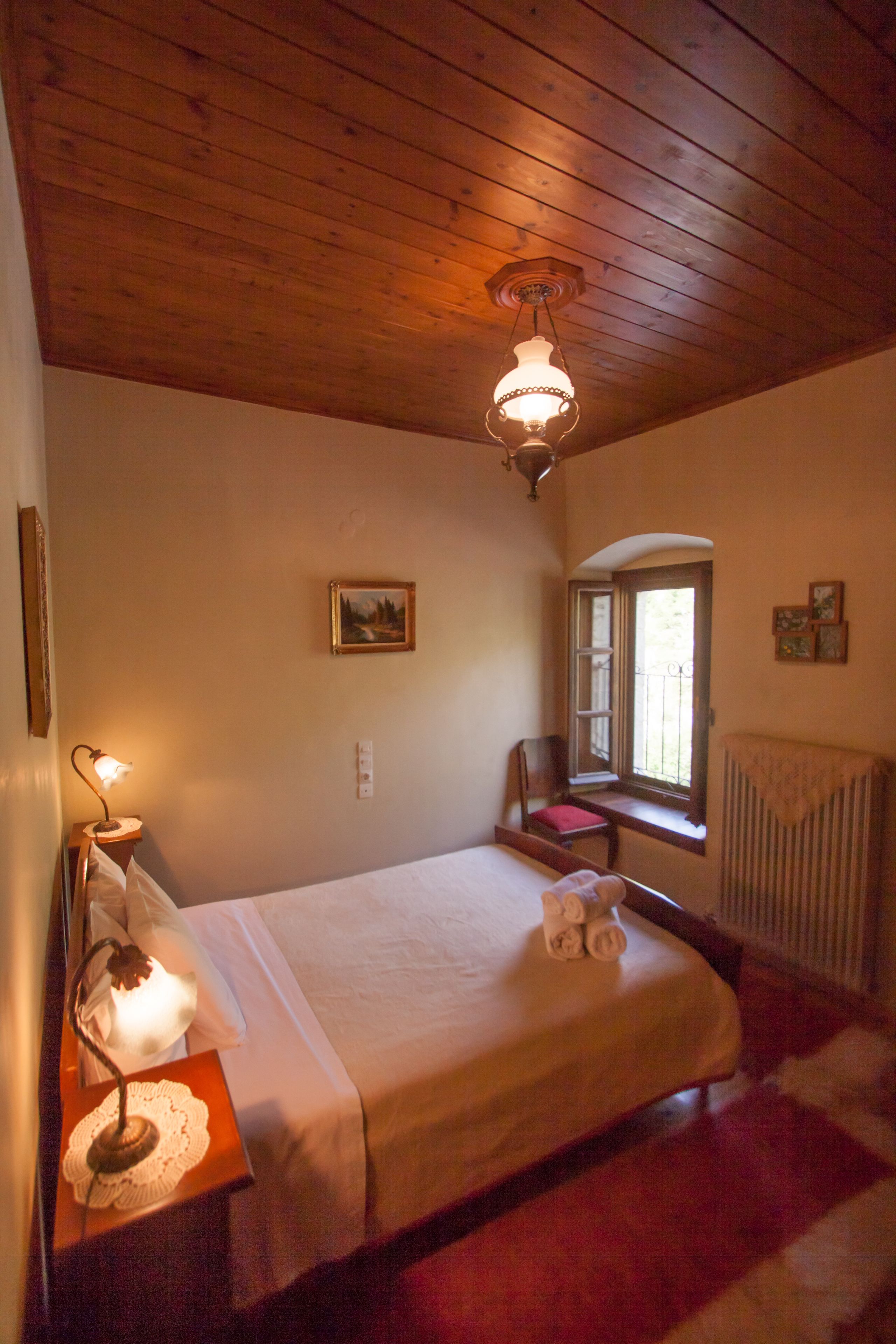 Une chambre avec un plafond en bois, un lit recouvert d’une couette blanche, une lampe de chevet, une fenêtre donnant sur un paysage agréable et un radiateur.
