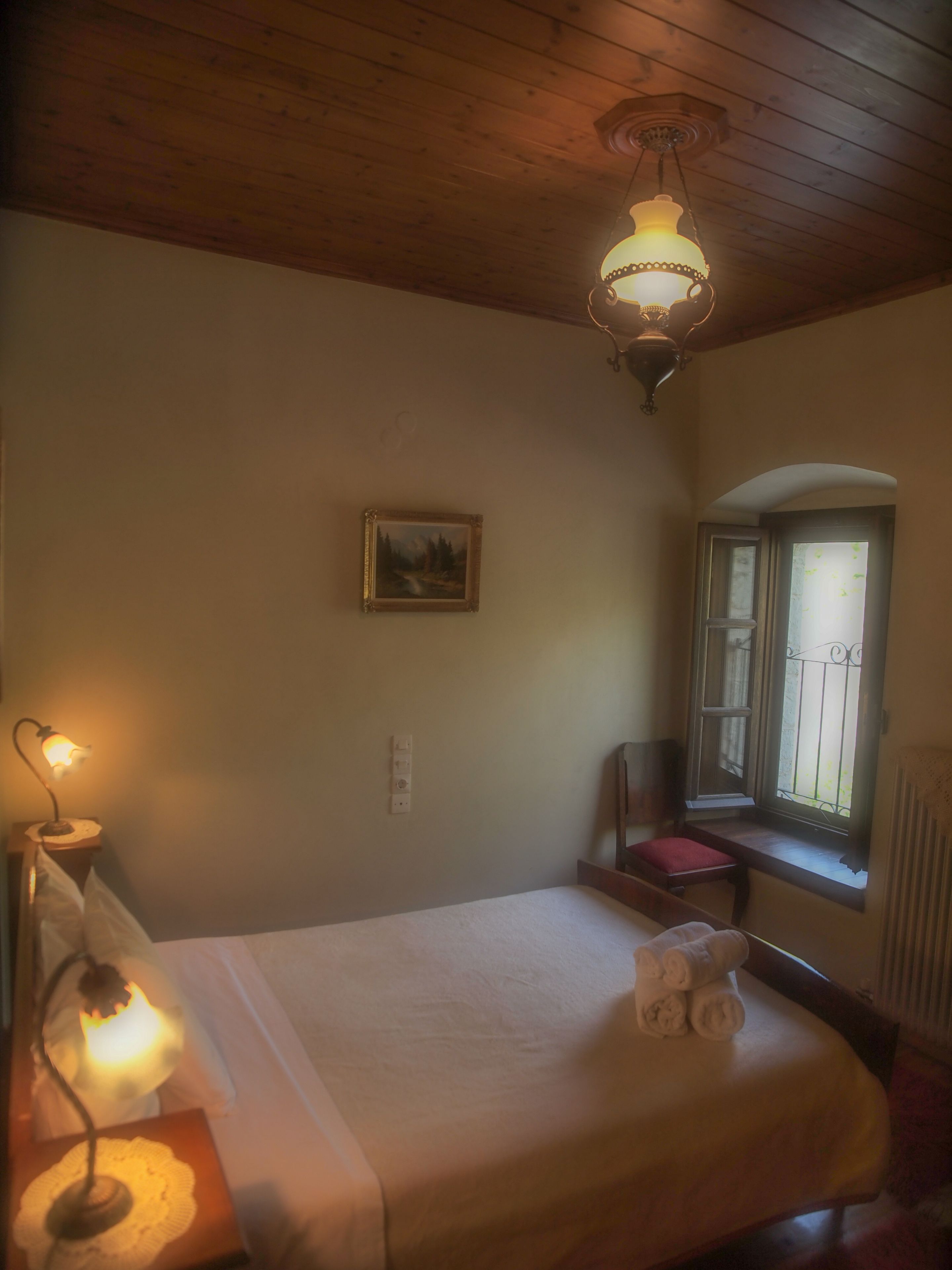 Une chambre avec un plafond en bois, un lit recouvert d’une courtepointe blanche, une lampe de chevet, un tableau encadré au mur et une fenêtre donnant sur un paysage verdoyant.