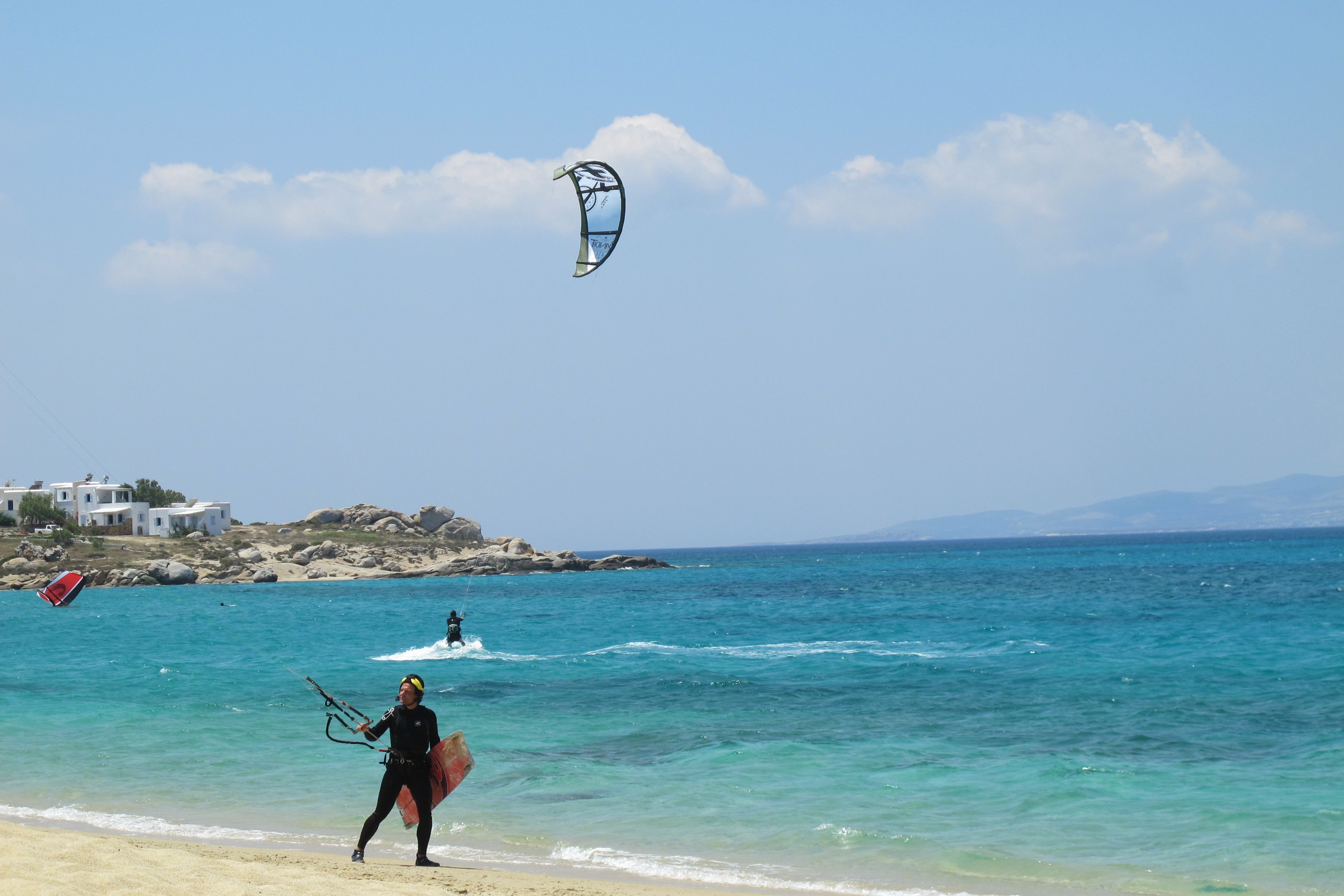 Una playa con agua azul cristalina, una persona en traje de neopreno sosteniendo una cometa para kitesurf, y un edificio al fondo.