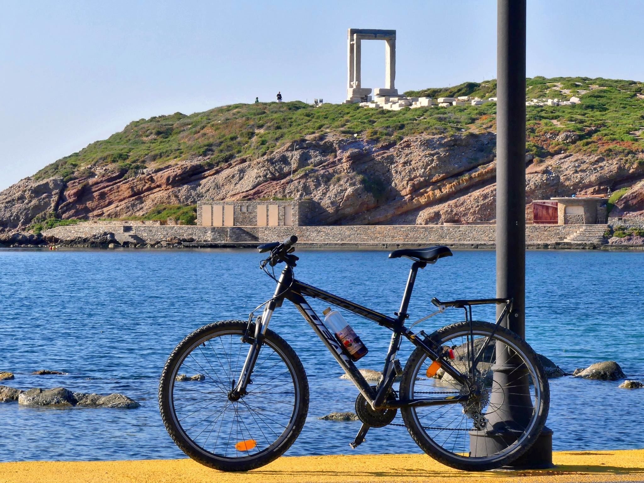 A bicycle parked near a waterfront with a rocky hill and a stone structure in the background.