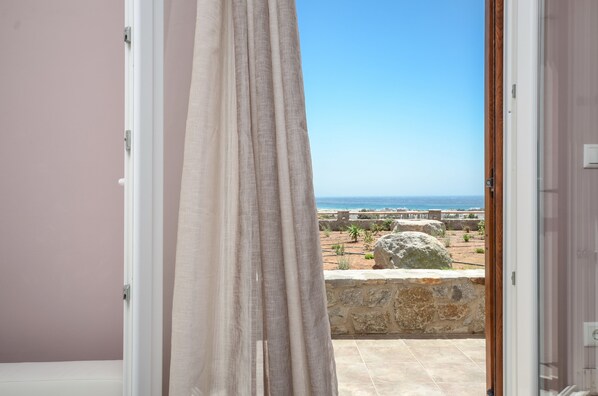 An open door leading to a balcony with a view of the sea and a stone wall.