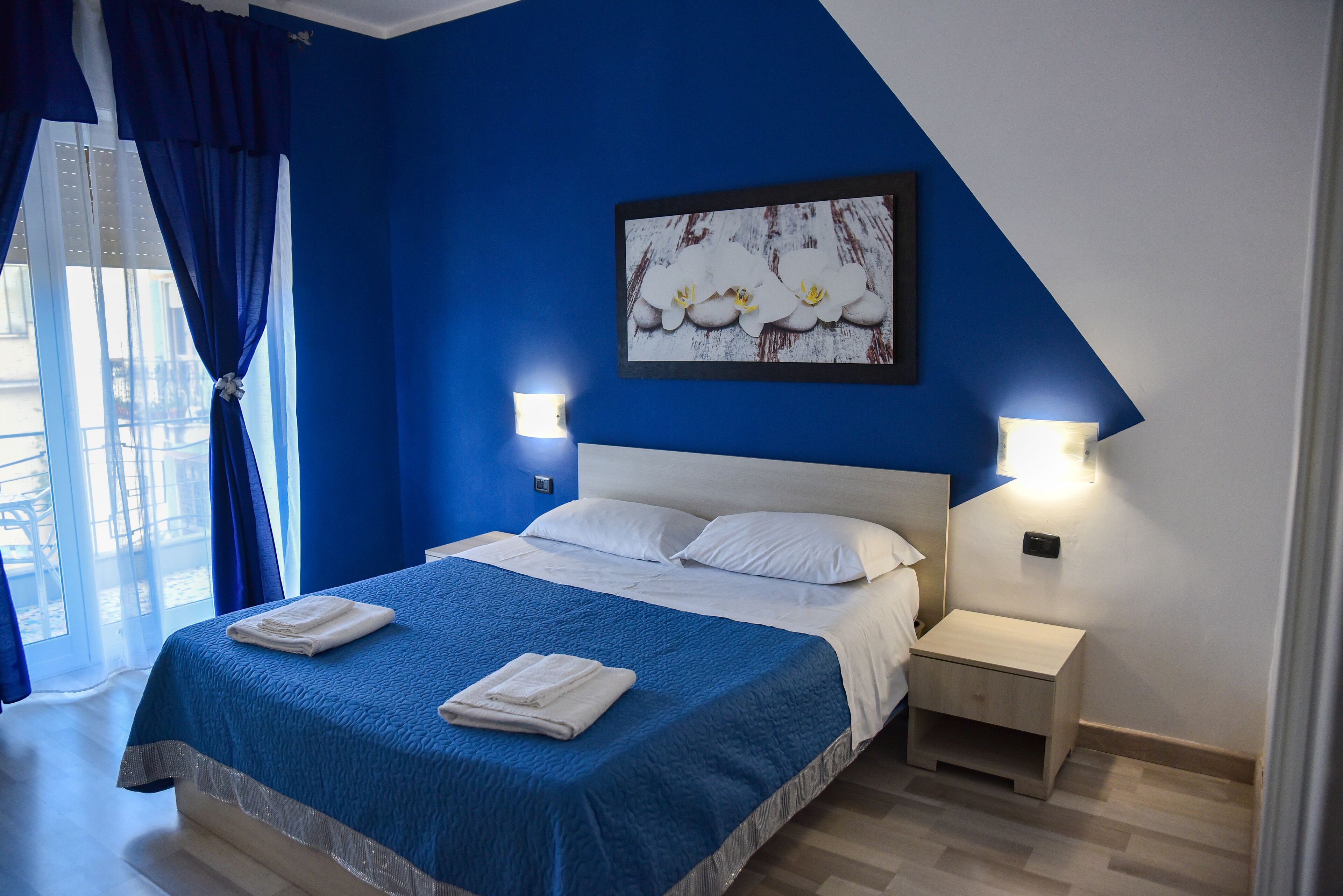 A bedroom with a blue wall, a bed with white bedding, a nightstand, and a painting of flowers.