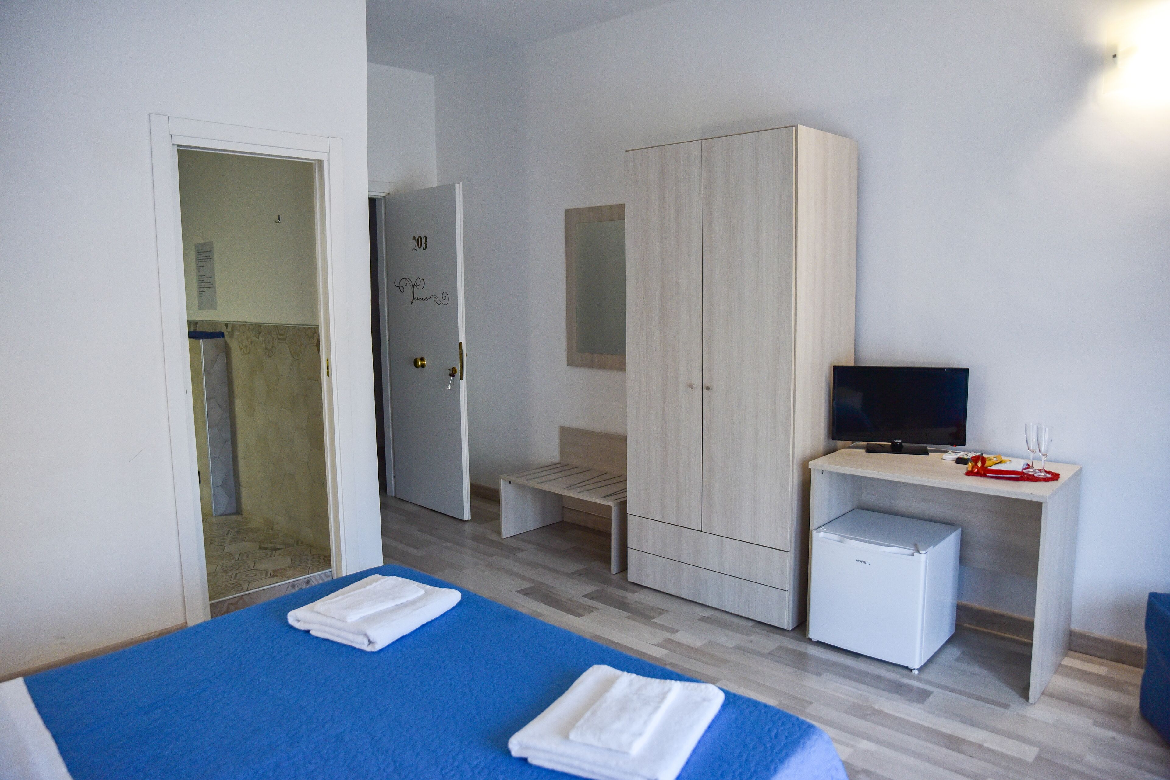 A bedroom with a bed, a desk with a TV, a wardrobe, and a small fridge.