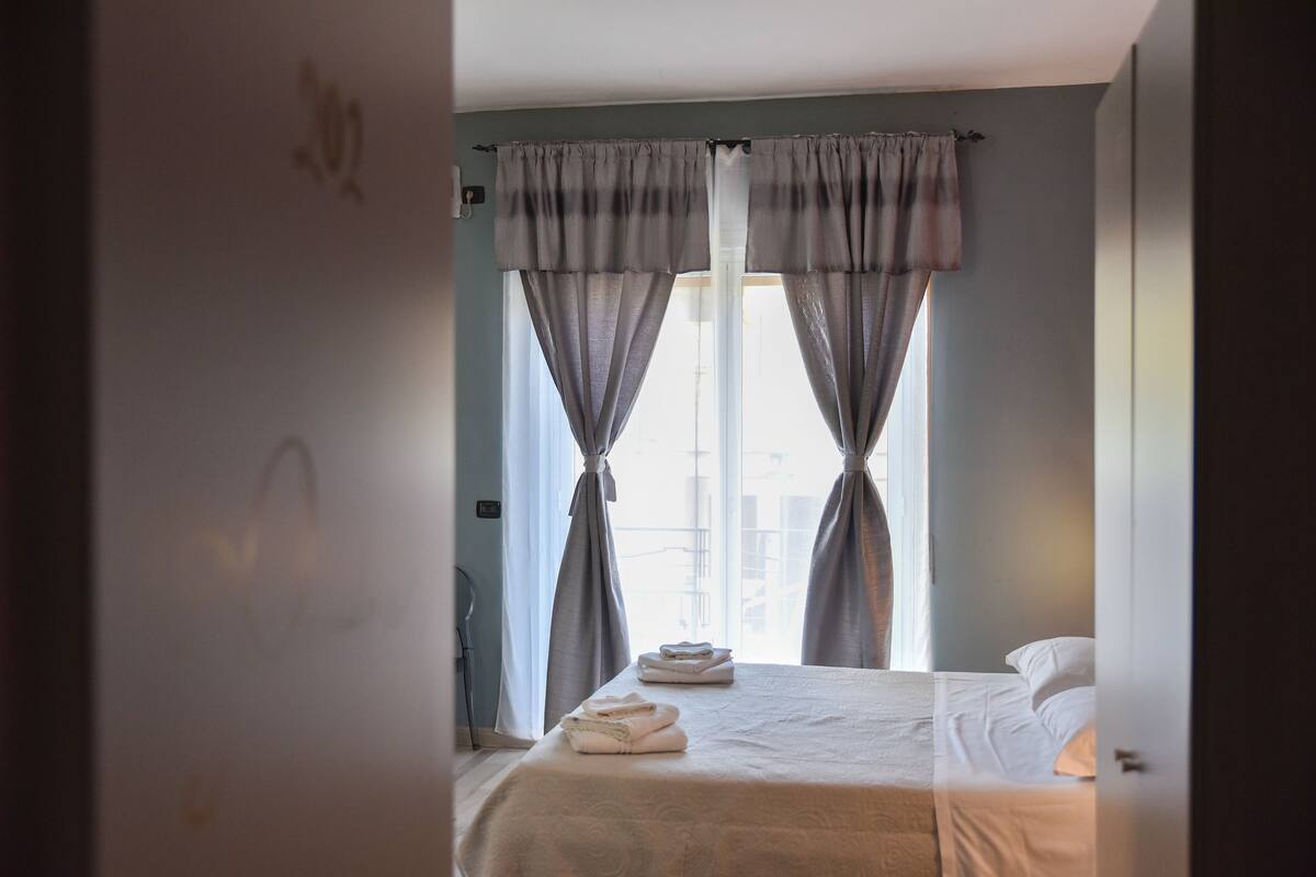 Quarto de hotel com cama, janela com cortinas e vista para a cidade.