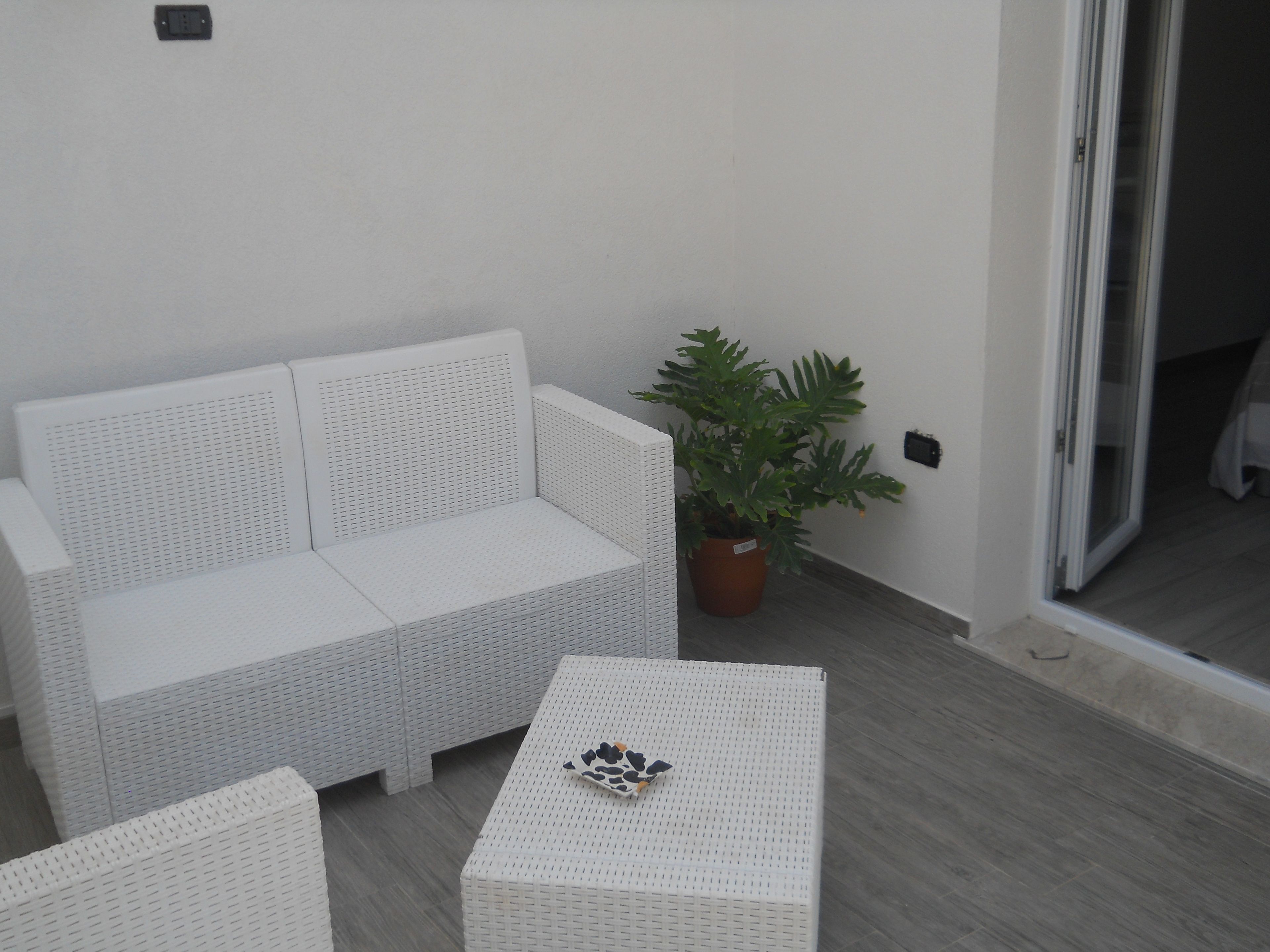 A white wicker sofa and two matching ottomans on a balcony with a potted plant.