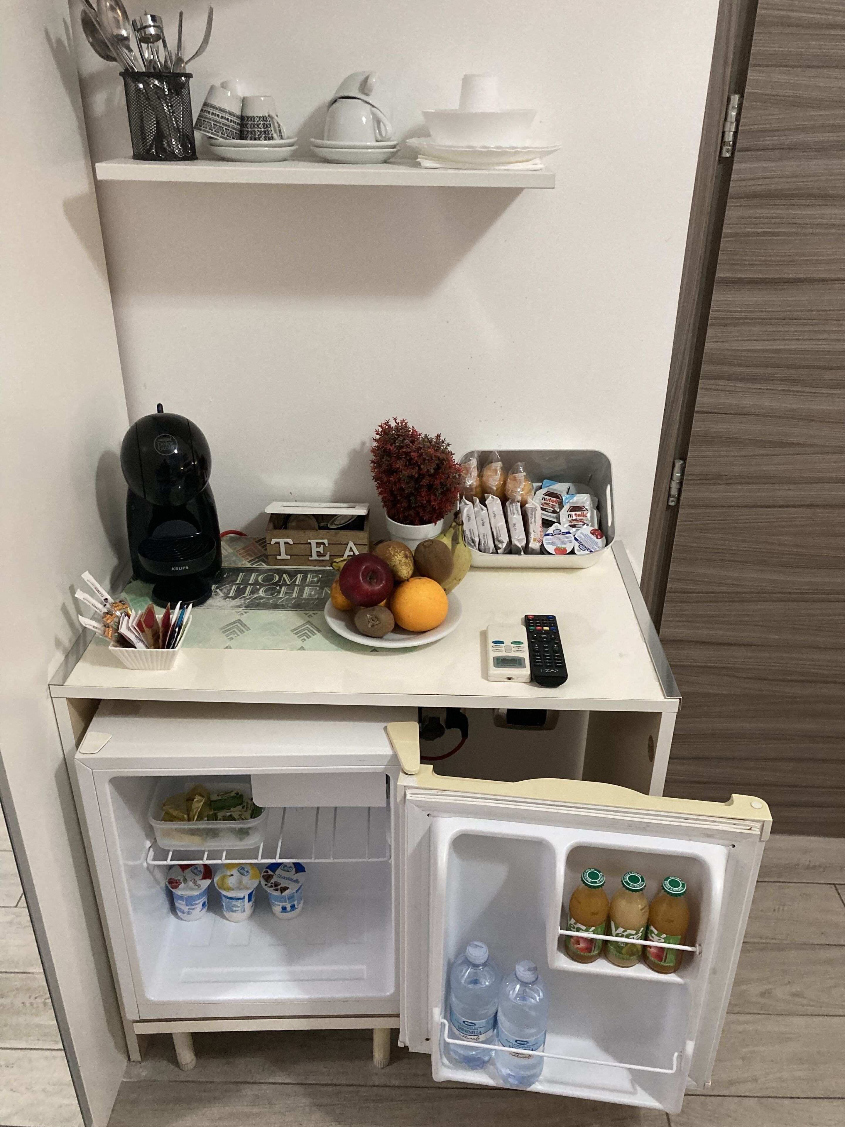 A compact kitchenette with a coffee maker, a bowl of fruit, a plant, and a fridge containing bottled water and juice.