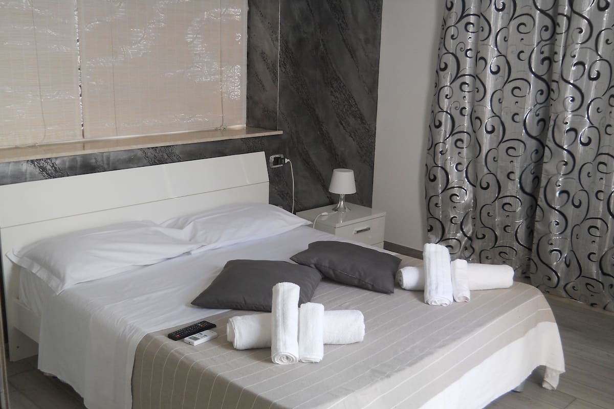 Una cama bien tendida con ropa de cama blanca y gris, un control remoto y toallas enrolladas sobre un piso de madera.
