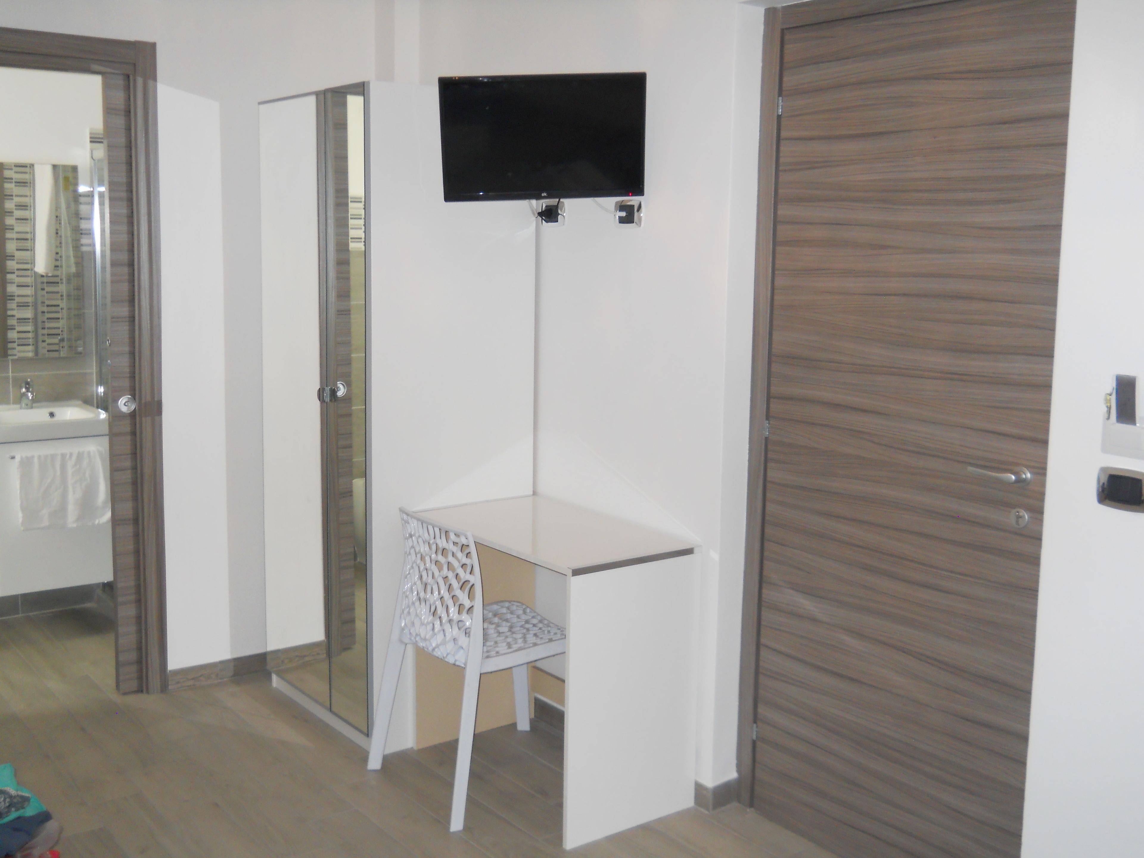 Ein Zimmer mit weißem Schreibtisch, einem Stuhl, einem an der Wand befestigten Fernseher, einer Holztür und einer Glastür zum Badezimmer.