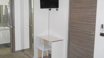 Ein Zimmer mit weißem Schreibtisch, einem Stuhl, einem an der Wand befestigten Fernseher, einer Holztür und einer Glastür zum Badezimmer.