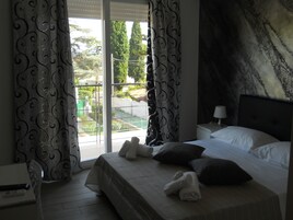 Habitación de hotel con cama, almohadas, mesita de noche, lámpara y vistas a un balcón con vegetación.