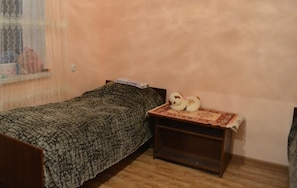 A small, single-bed room with a patterned bedspread, a bedside table with a teddy bear, and a window with a sheer curtain.