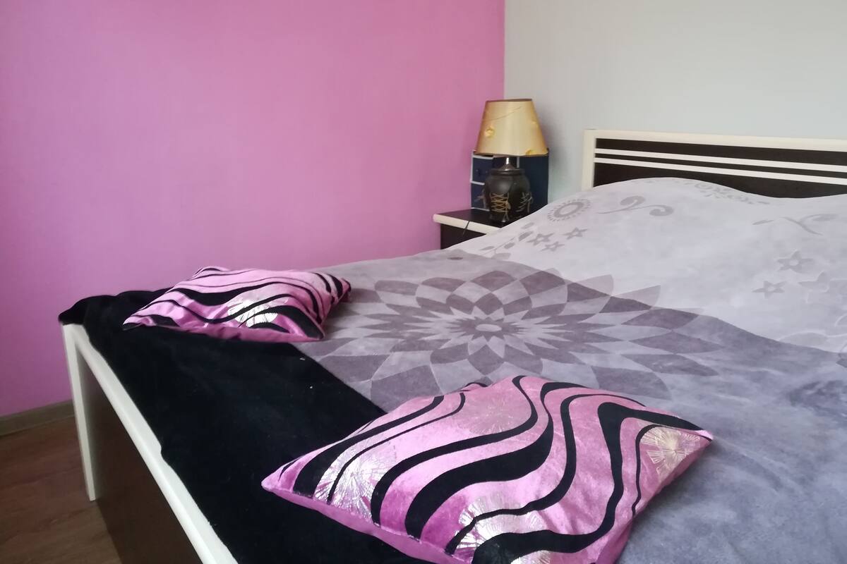 Una cama con una colcha estampada y dos almohadones decorativos, una lámpara de noche y una mesita de noche con lámpara.