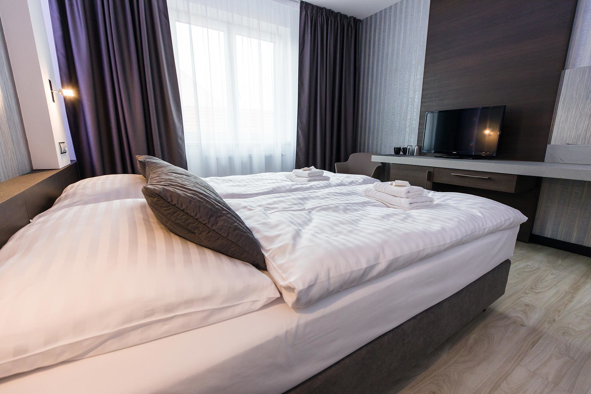 Moderni hotellihuone, jossa on suuri sänky, työpöytä ja litteänäyttöinen televisio sekä suuret ikkunat, joista avautuu näkymä kaupunkiin.