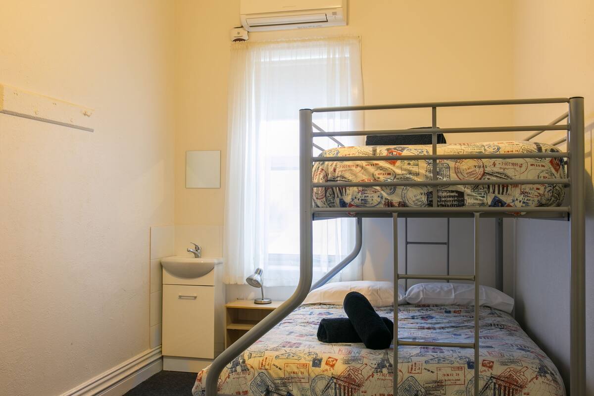 A bunk bed with a patterned bedspread, a bedside table with a lamp, a small white cabinet, and a window with sheer curtains.