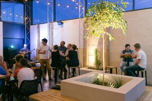 Un área de comedor al aire libre con mesas y sillas, un escenario con un guitarrista, y un árbol grande.