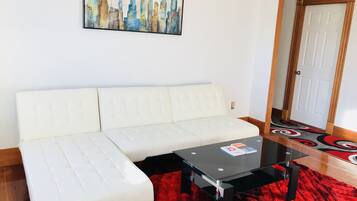 Um sofá branco, uma mesa de centro de vidro e um tapete vermelho em um quarto com uma pintura de paisagem urbana na parede.