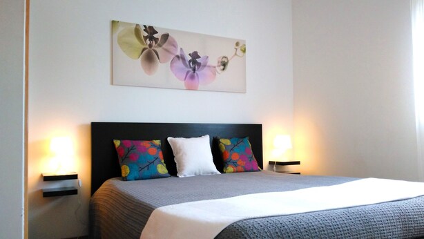 Ein ordentlich bezogenes Bett mit grauer Decke, zwei Kissen und einem floralen Kunstwerk darüber. Auf jeder Seite des Bettes befinden sich Nachttische mit Lampen.
