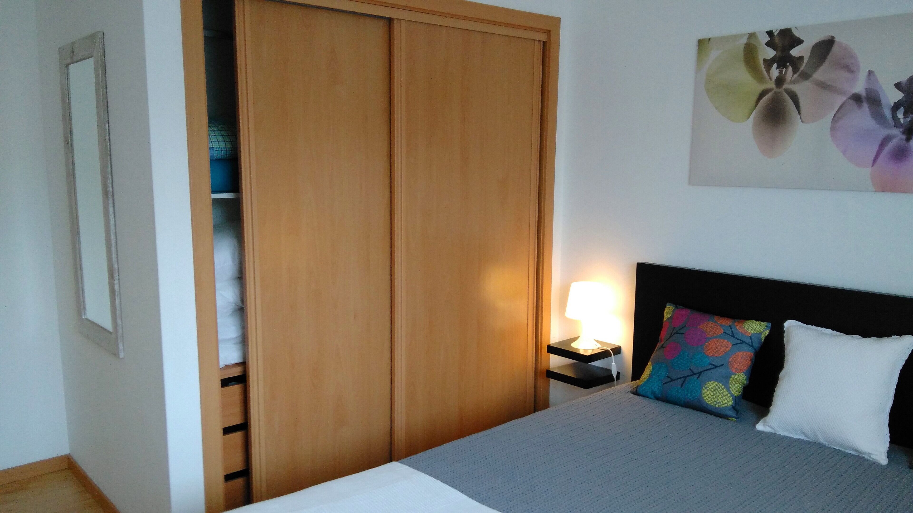 A bedroom with a bed, a nightstand, a lamp, a wardrobe, and a wall-mounted artwork.