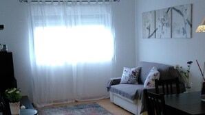 Ein Wohnzimmer mit Sofa, Esstisch, Couchtisch und einem Fenster mit transparenten Vorhängen.