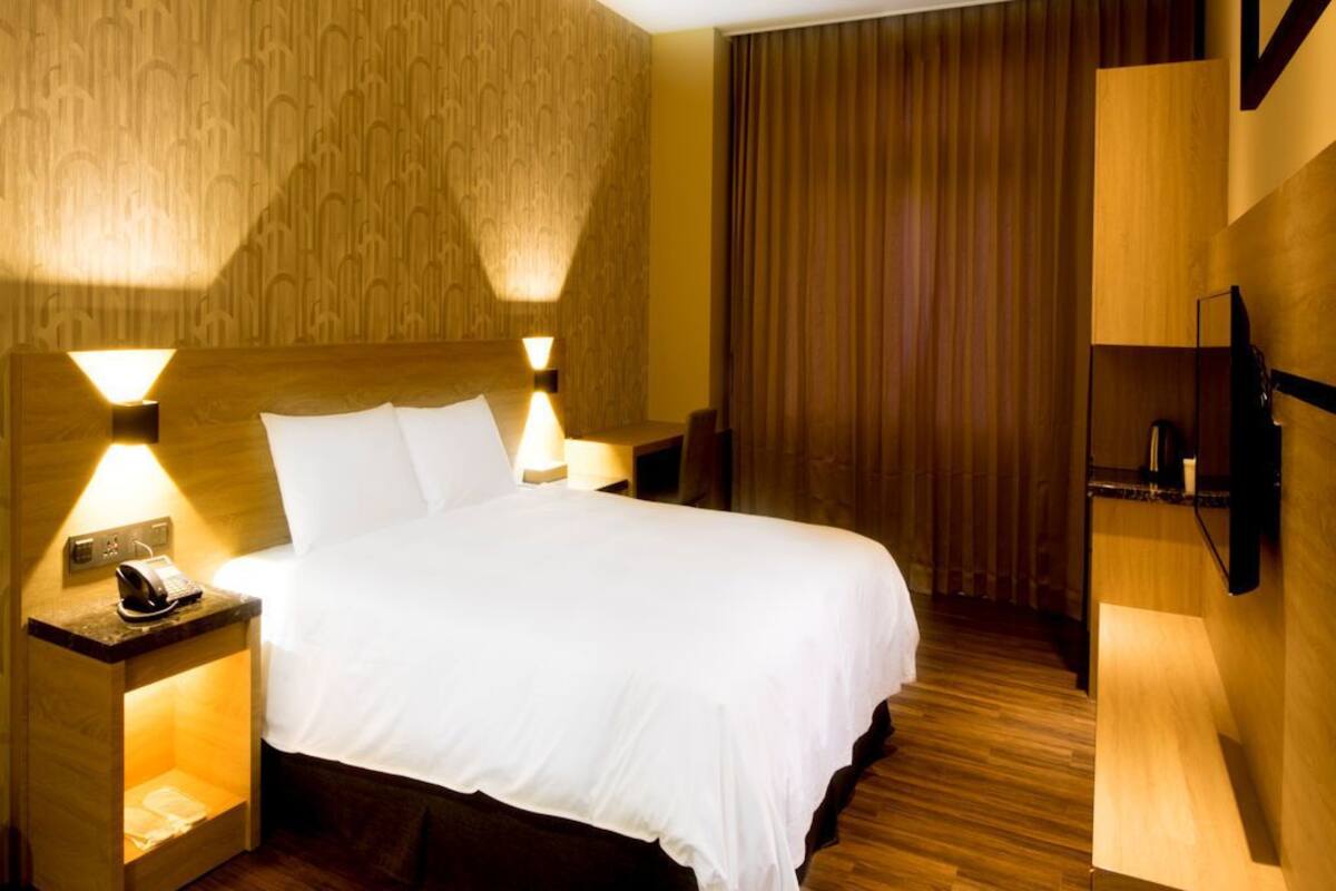 A hotel room with a wooden headboard, a bed with white linens, two bedside lamps, a nightstand with a phone, and a flat-screen TV mounted on the wall.