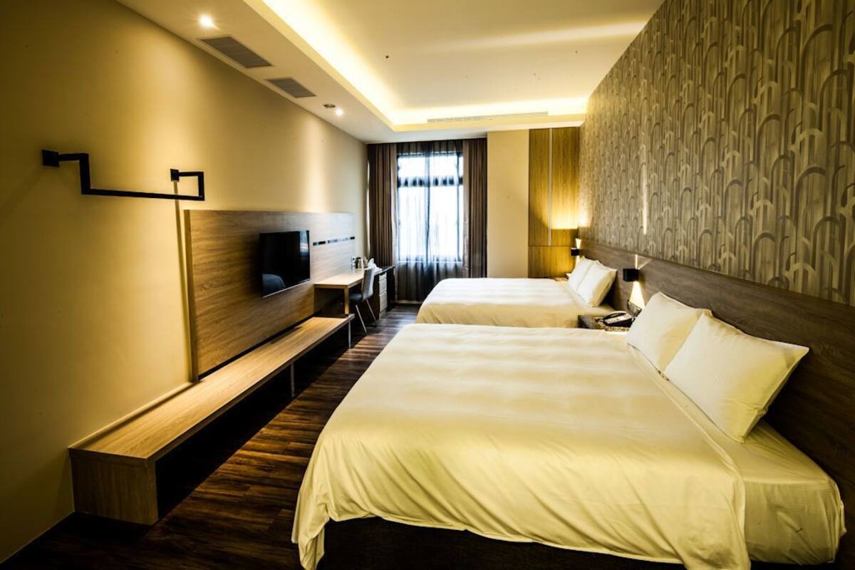 A hotel room with two beds, a desk, a TV, and a patterned wall.