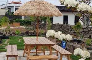 A wooden picnic table with benches and a thatched umbrella in a garden area with potted plants and a stone wall. - HANI Haus (Jeju City)