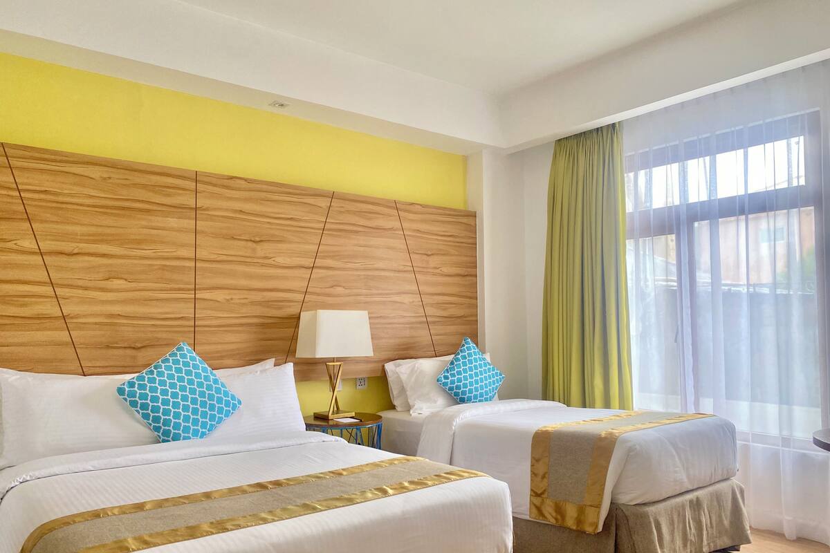 Ein Hotelzimmer mit zwei Betten, einem Holzkopfstück, einem Nachttisch mit Lampe und einem Fenster mit Vorhängen.