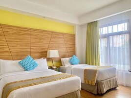 A hotel room with two beds, a wooden headboard, a nightstand with a lamp, and a window with curtains.