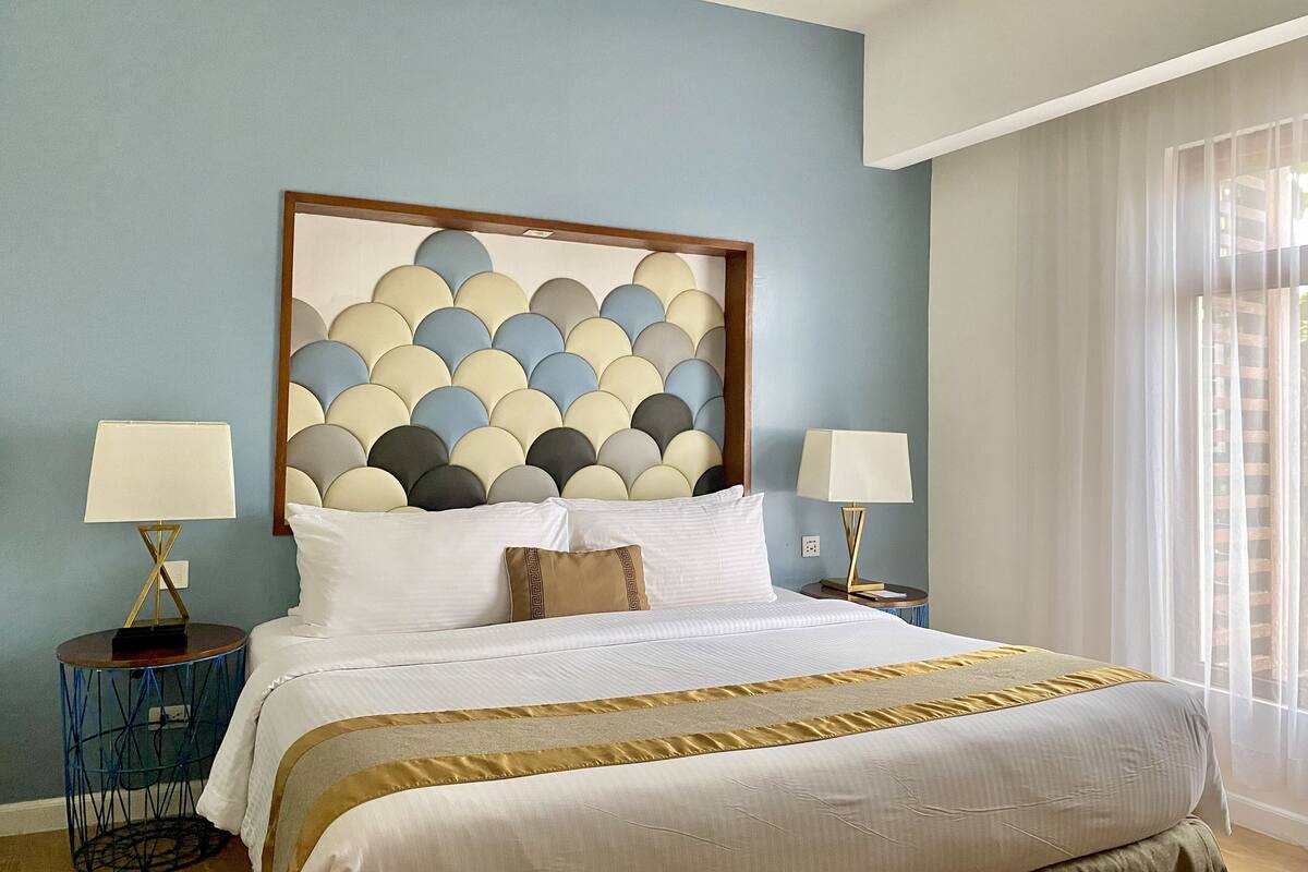 Ein Hotelzimmer mit einem Bett, zwei Nachttischen, einer Wand mit geometrischem Muster und einem Fenster mit transparenten Vorhängen.